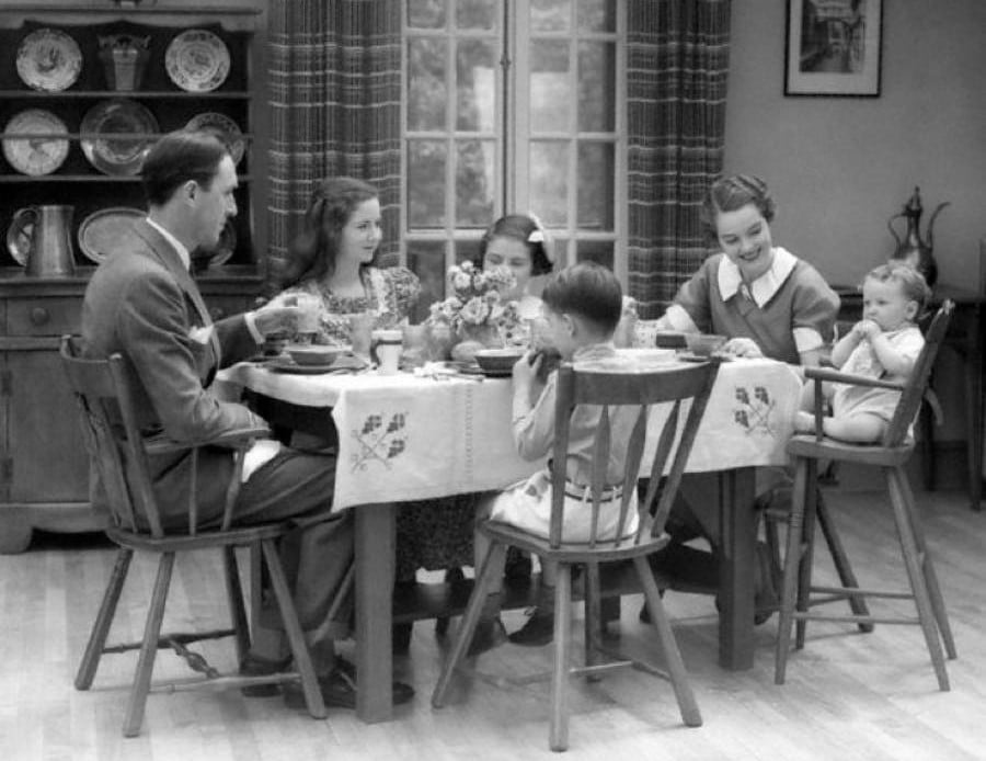 A black-and-white photo of a family of six, including two adults and four children, sitting around a dining table, sharing a meal in a cozy, vintage-style kitchen or dining room.