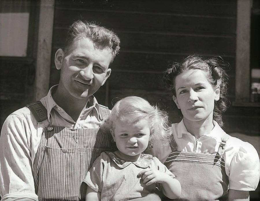 A black-and-white photo of a man and woman in overalls standing close together, with a small child between them. The man and child are smiling, and the woman has a neutral expression. They are outdoors in front of a dark building.