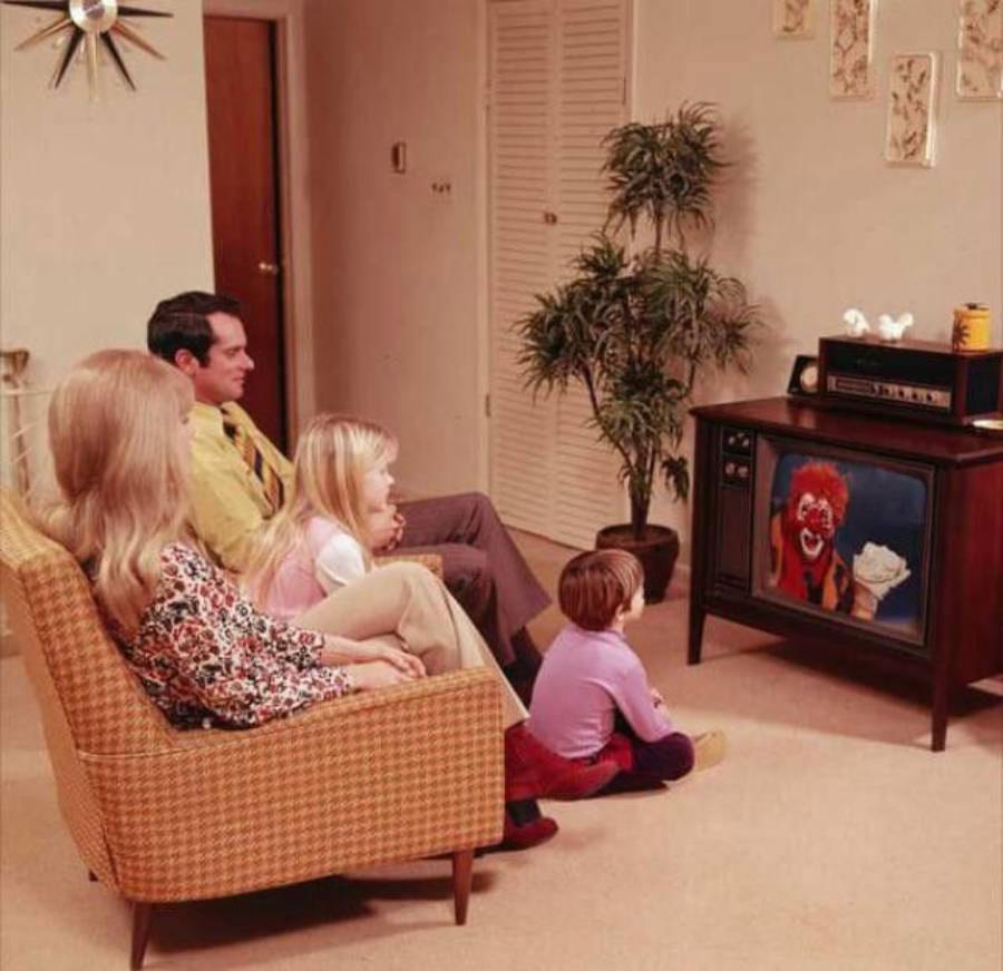 A family of four sits on a couch and floor in a living room, watching a TV showing a clown. The room has beige walls, plants, and 1970s-style decor.