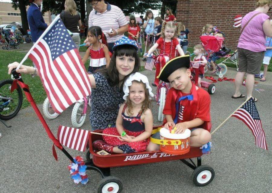 Three children in patriotic costumes sit in a red wagon with American flags during a parade. Other kids and adults are in the background, many with decorated bicycles, celebrating outdoors on a sunny day.