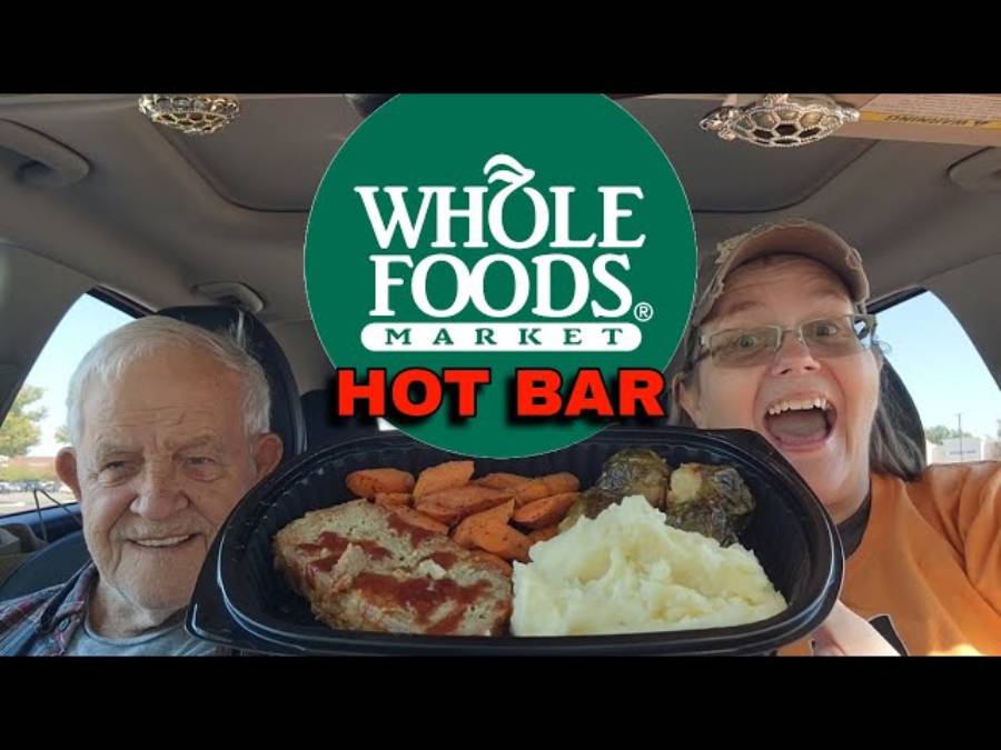 Two people smile in a car holding a tray of hot food. The Whole Foods Market logo and the words "HOT BAR" are displayed above the tray.