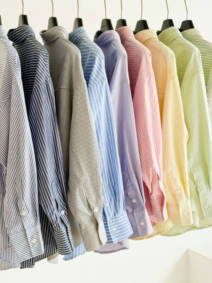 Striped button-down shirts in various pastel colors hang neatly on black hangers in a row, ranging from navy, gray, and blue to purple, pink, peach, yellow, and green.