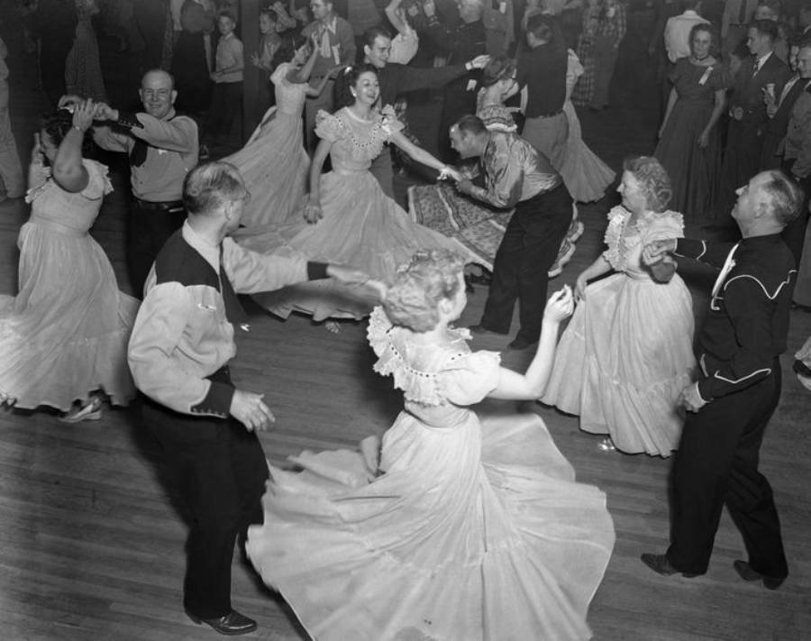 Men and women in vintage attire joyfully square dance in a crowded ballroom, twirling and holding hands, with full skirts swirling around them on a wooden floor.