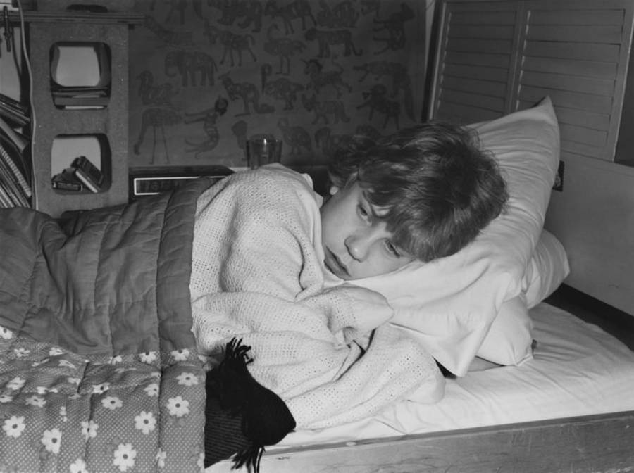 A child with light hair is lying in bed on their side, resting their head on a pillow, partially covered by a blanket, and looking away thoughtfully. The room has animal drawings on the wall and books on a shelf.