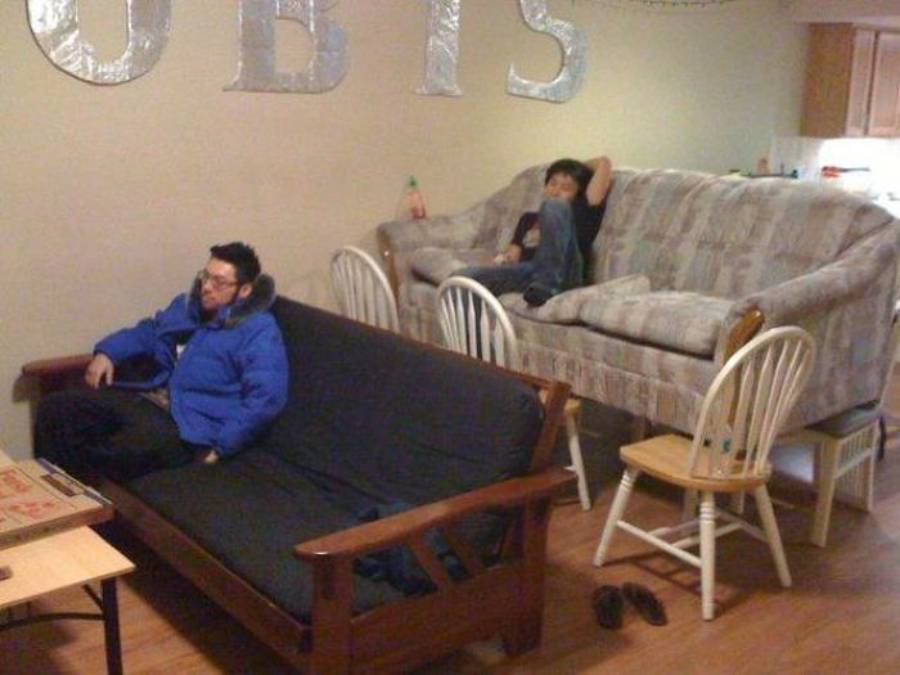 Two people watch TV; one sits on a futon while the other sits on a couch that is elevated by four chairs. A small table and shoes are in the foreground, and large silver letters hang on the wall in the background.