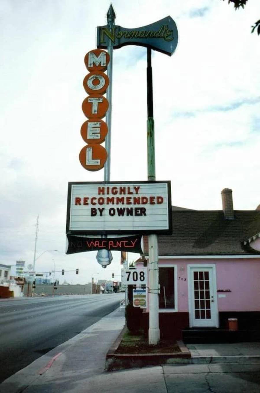A vintage motel sign reads "Normandie Motel" with a marquee below stating "Highly Recommended by Owner." A "No Vacancy" sign and address number 708 are also visible outside a pink building by the roadside.