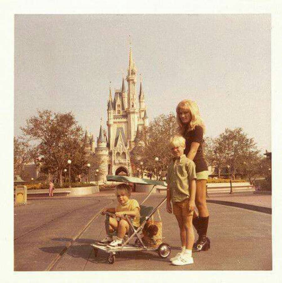 Three children pose in front of Cinderella Castle at Disney World. One sits in a stroller, another stands beside, and the oldest stands behind them. The scene suggests a vintage photo from the 1970s.