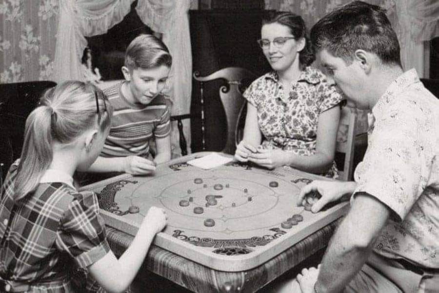 A black and white photo of four people, two adults and two children, sitting around a table playing a board game together in a cozy, vintage living room.