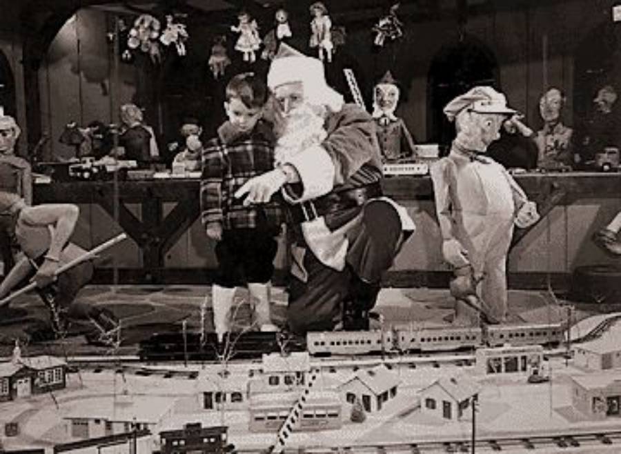 Santa and a boy look at a model train display, surrounded by elves and toys in a festive, vintage holiday scene.