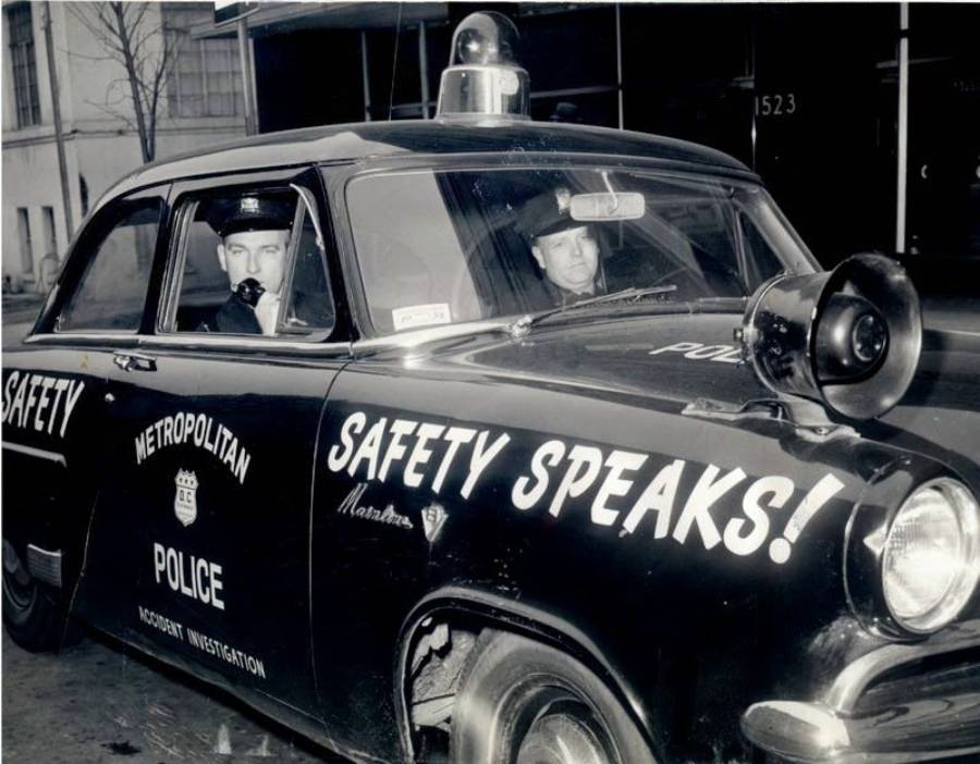 Two police officers sit in a vintage Metropolitan Police car with “SAFETY SPEAKS!” painted on the door and a large loudspeaker mounted on the hood. The car is parked on a city street.