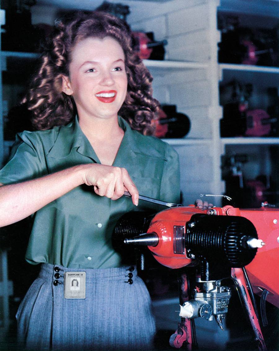 A smiling woman with curly hair in a green blouse uses a wrench on a red machine in a workshop. She wears a skirt with an ID badge clipped to it. Shelves with equipment are visible in the background.