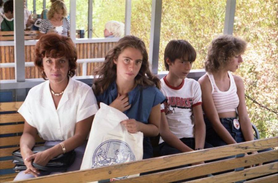 Four people sit on a wooden bench outdoors. Two women are on the left, and two teenagers, a boy and a girl, are on the right. They all appear serious or deep in thought. Trees and other people are visible in the background.