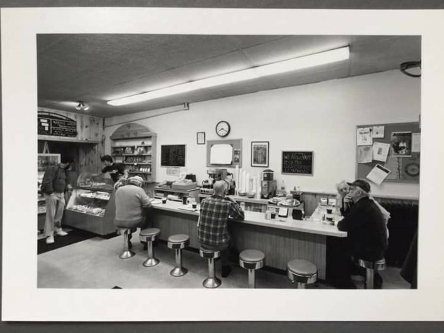 Black and white photo of a diner with a counter, five people seated on stools, and a display case. Two people stand near the counter. Menus and notices are on the wall behind, along with a clock and coffee machines.