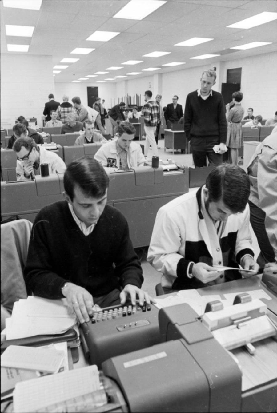 Black-and-white photo of a busy office or classroom in the 1960s, with people working at desks using vintage adding machines and paperwork. Several people stand or walk in the background.