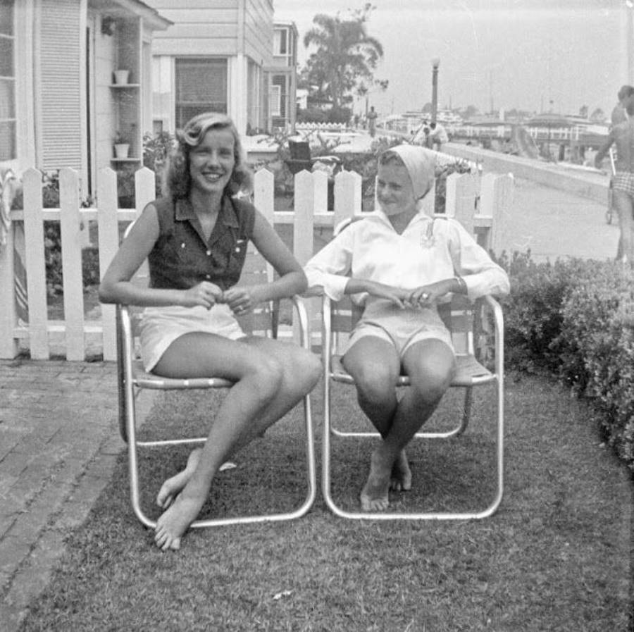Two women sit barefoot on metal lawn chairs in a yard, smiling, with a white picket fence and houses in the background. A canal and another person walking are visible in the distance.