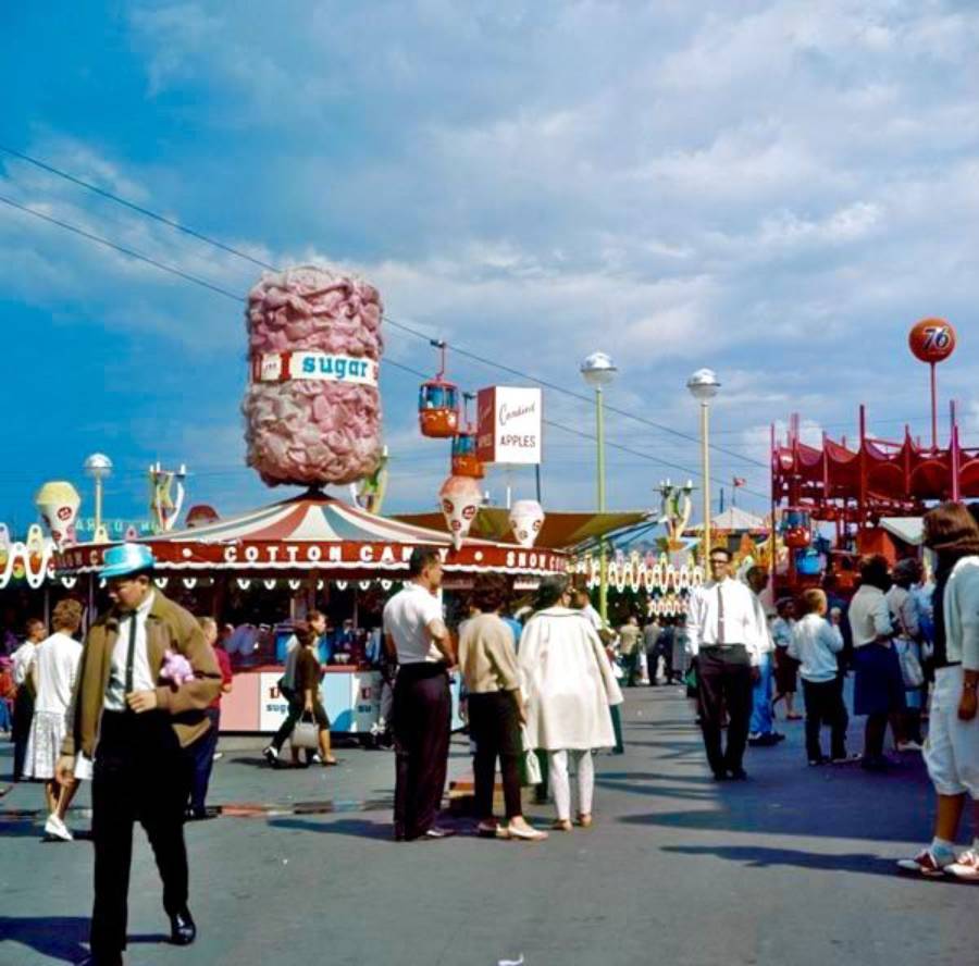 Colorful fairground scene with crowds of people walking near a large cotton candy stand, carnival games, and rides, including a gondola lift in the background under a partly cloudy sky.