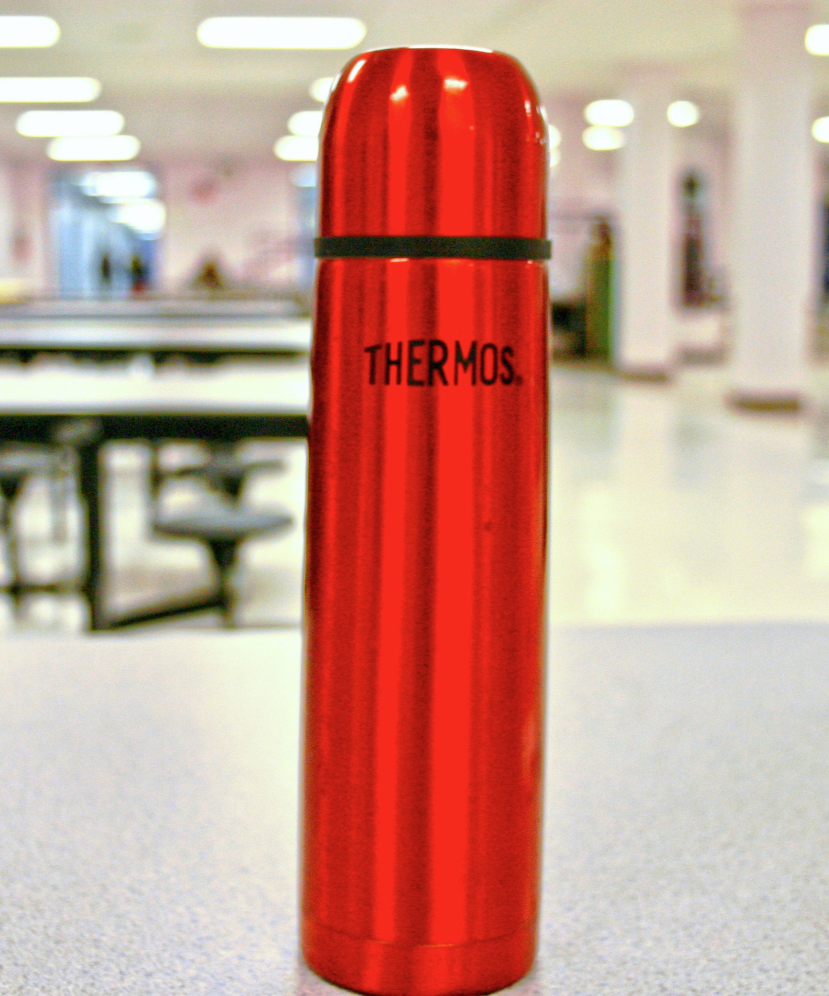 A shiny red thermos stands upright on a table in a brightly lit room with blurred tables and chairs in the background.
