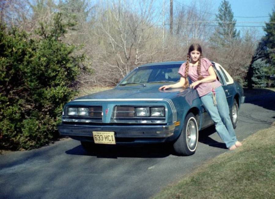 A young woman with long brown hair, wearing jeans and a pink top, leans against the front of a blue vintage car parked on a driveway, surrounded by trees and bushes.