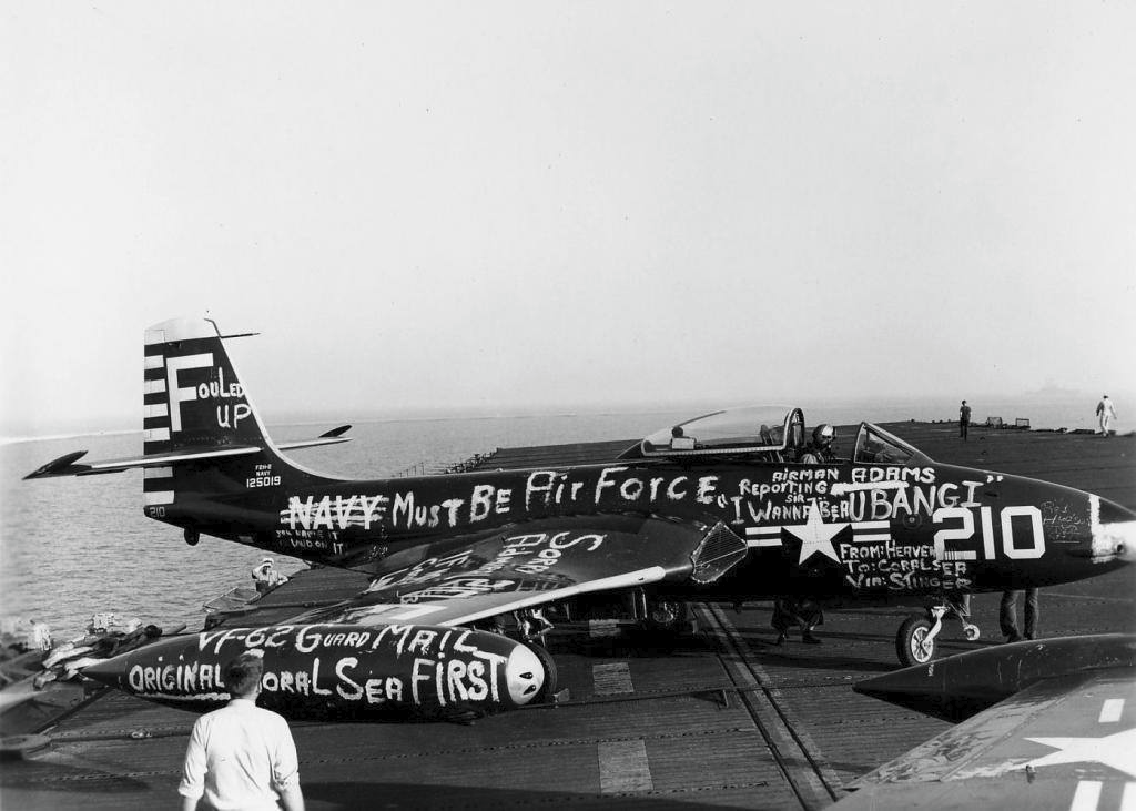 A military aircraft on a carrier deck covered in graffiti-like chalk messages, including “Navy must be air force,” “Bang!,” and “From heaven to carrier via Smiser.” People stand nearby.