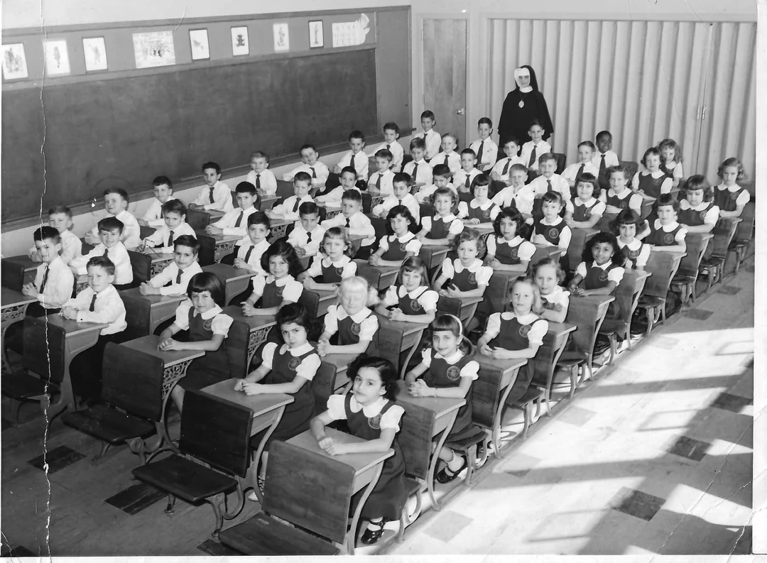 Black and white photo of a classroom filled with young girls in uniform sitting at desks, with a nun standing at the back. The classroom has a chalkboard, drawings displayed, and sunlight streaming through windows.