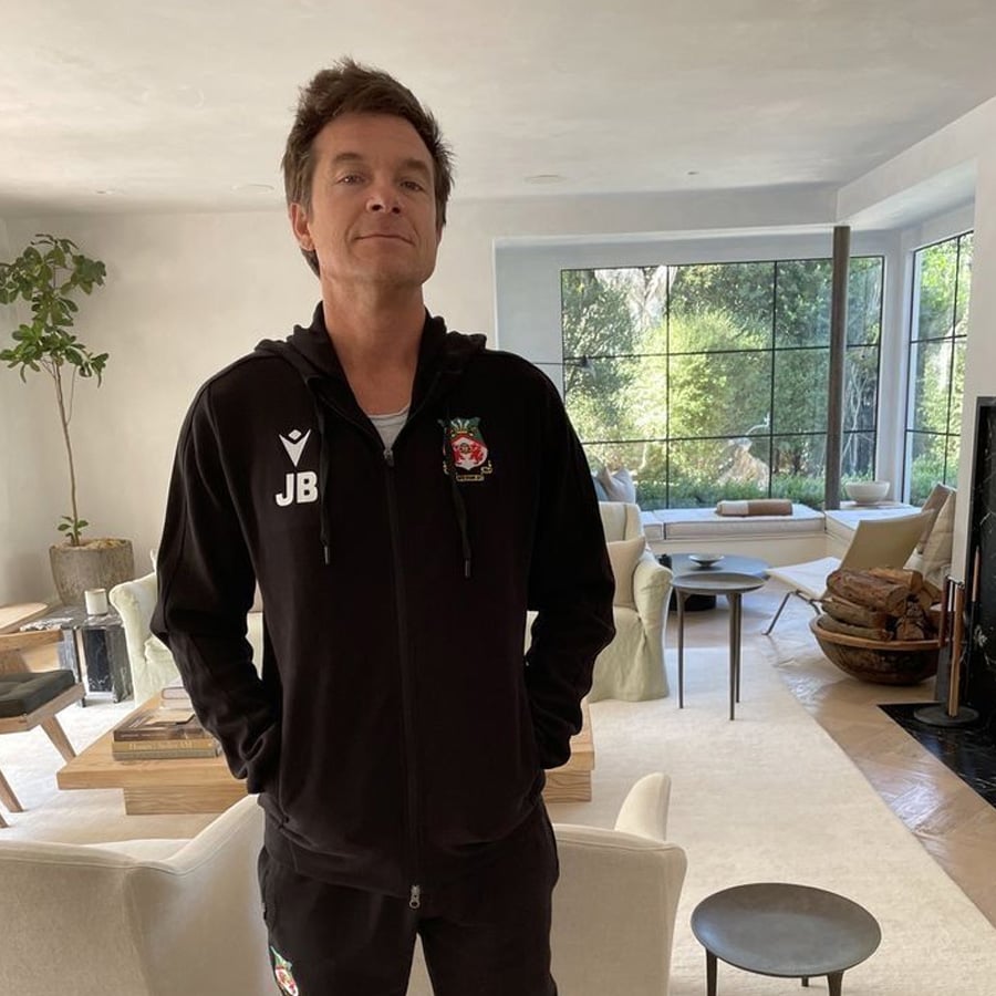 A man with brown hair stands in a modern, bright living room wearing a black zip-up jacket with "JB" and a red emblem on it. Large windows overlook a green garden in the background.