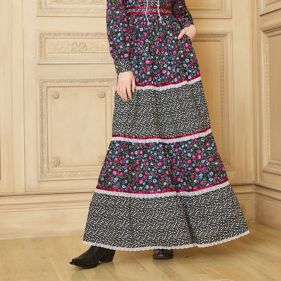 A person stands indoors wearing a long, floral-patterned dress with black, pink, blue, and white colors, and black shoes, against a beige paneled wall.