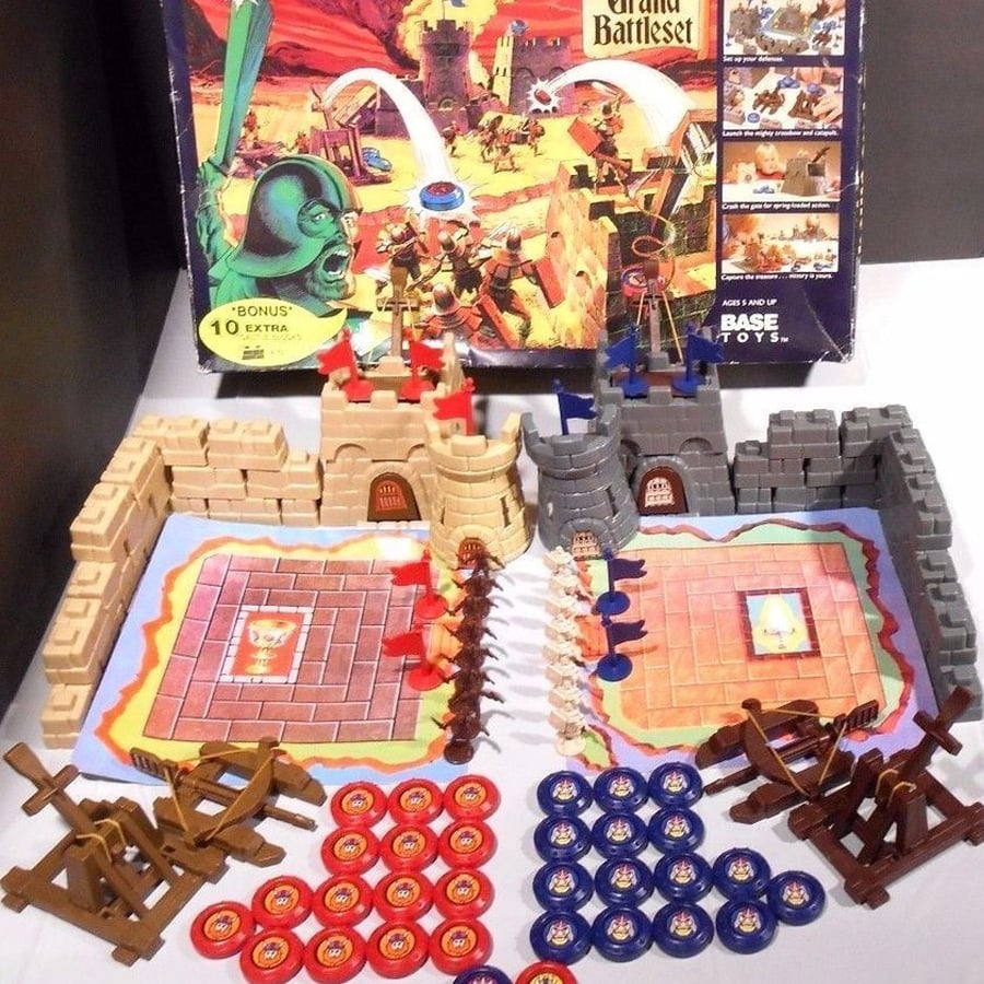 Two toy castle playsets with red and blue soldiers, catapults, coins, and castle walls, set up on illustrated mats. The game box with colorful battle artwork stands behind the playsets.