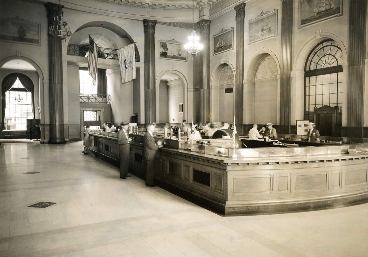 Historic black-and-white photo of a large, elegant bank lobby with high ceilings, chandeliers, marble floors, and several people standing or working at a long, wooden counter.