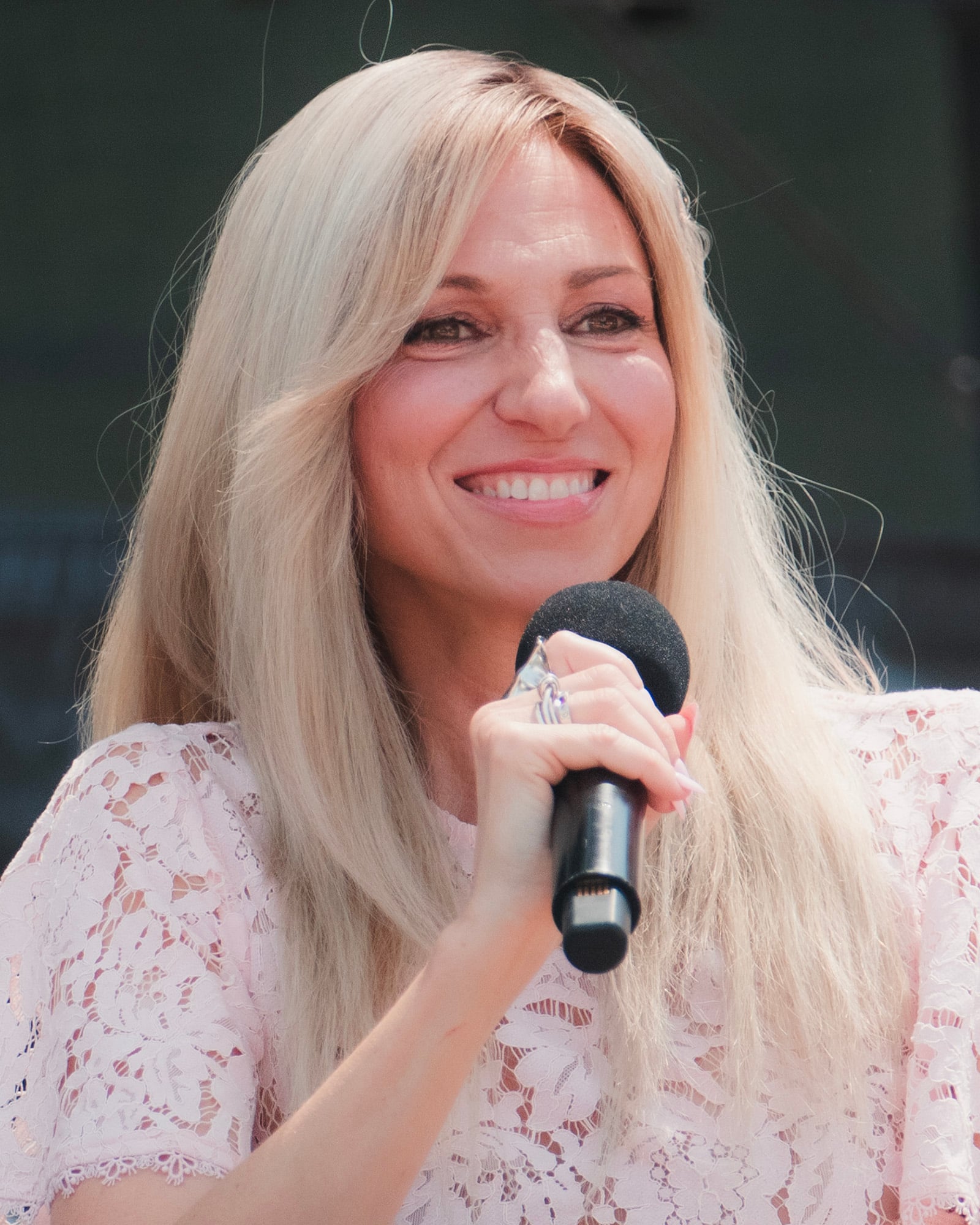 A woman with long blonde hair, wearing a white lace top, smiles while holding a black microphone.