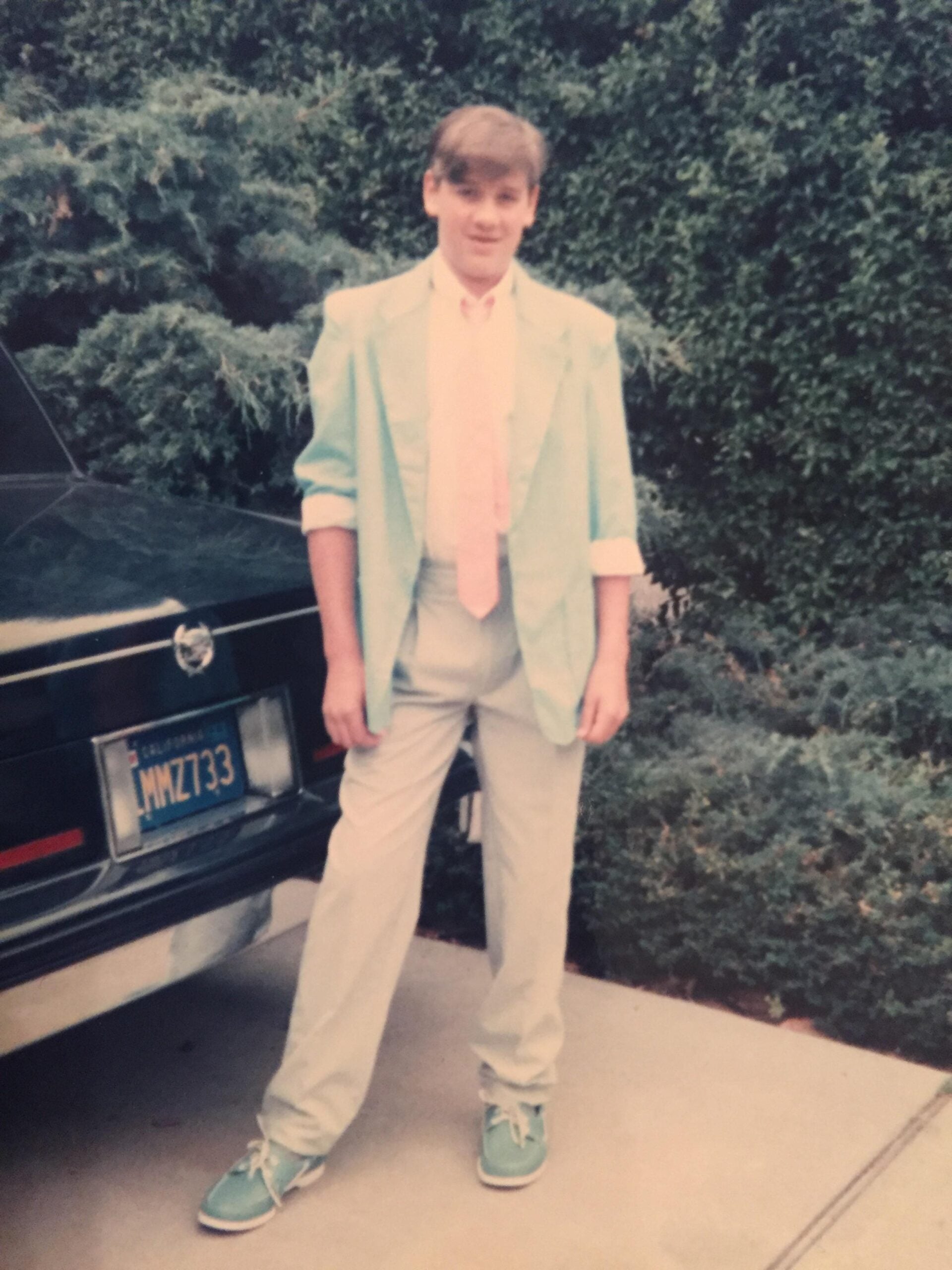 A young person stands outside by a car and bushes, wearing a pastel suit, pink tie, and matching teal shoes, smiling at the camera.