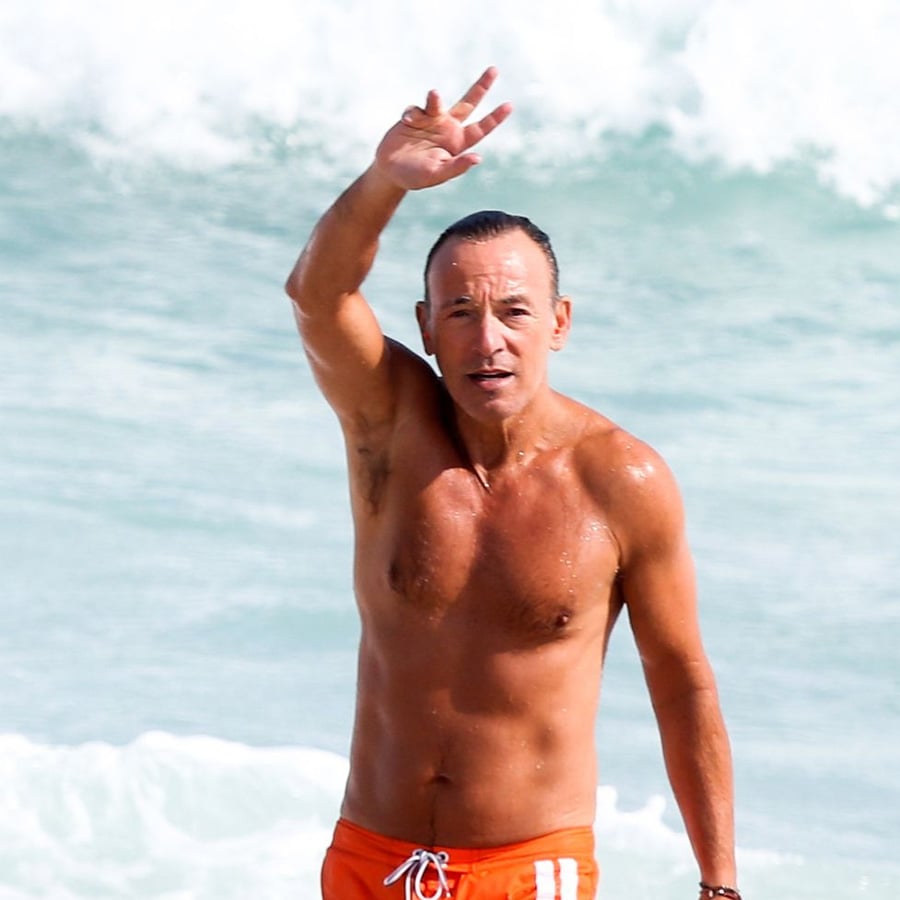 A man wearing orange swim trunks stands in shallow ocean water, waving with one hand. Waves are visible in the background.