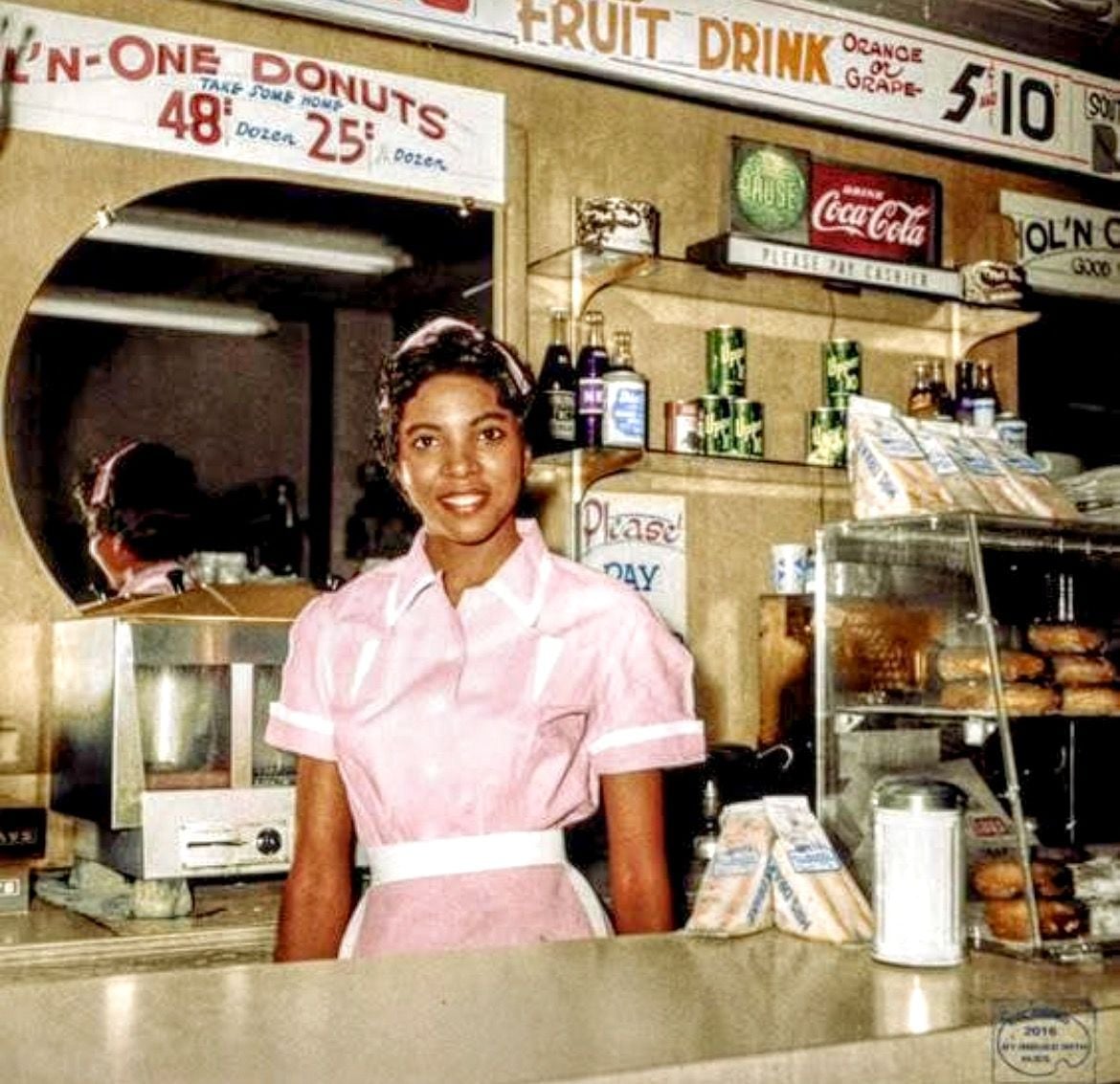 A smiling woman in a pink waitress uniform stands behind the counter of a retro diner with donuts, sandwiches, a Coca-Cola sign, and a menu displayed on the wall.