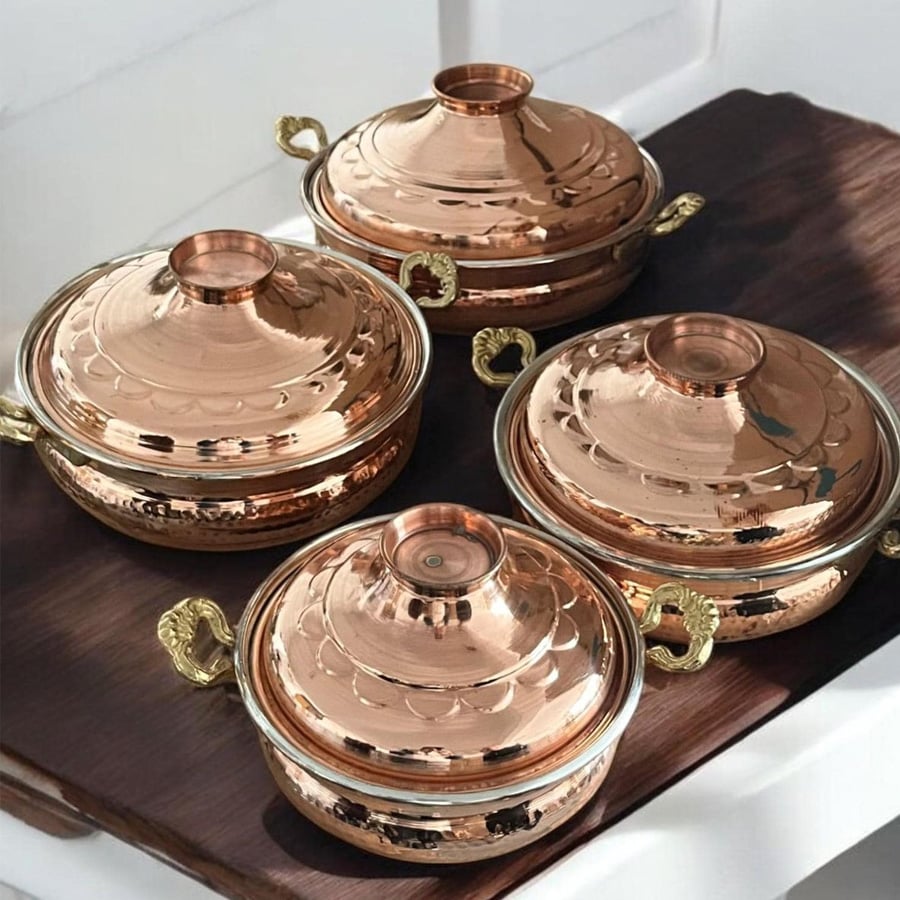 Four shiny copper pots with lids and decorative gold handles are arranged on a dark wooden surface, reflecting light.