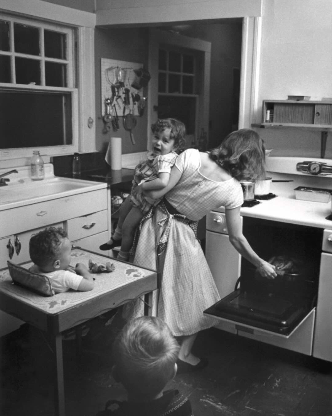 A woman holding a child opens an oven in a vintage kitchen. A toddler sits at a small table, while another child stands nearby, watching. The kitchen has a classic 1950s style.