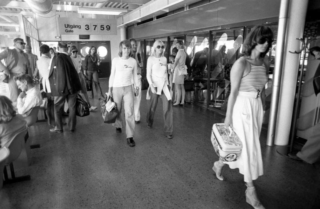 Black-and-white photo of people walking through an airport terminal. Most are dressed in 1970s fashion. Signs above indicate gates 3, 7, 5, and 9, and some people are seated on benches along the wall.
