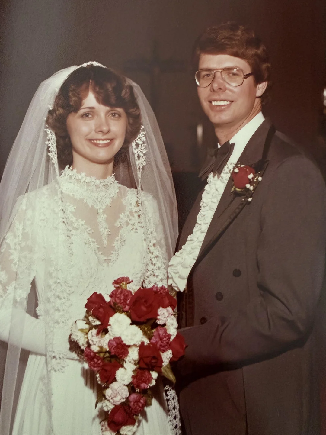 A bride in a lace wedding dress and veil stands next to a groom in a gray tuxedo with a bow tie and glasses. They are smiling and holding a bouquet of red and white flowers.