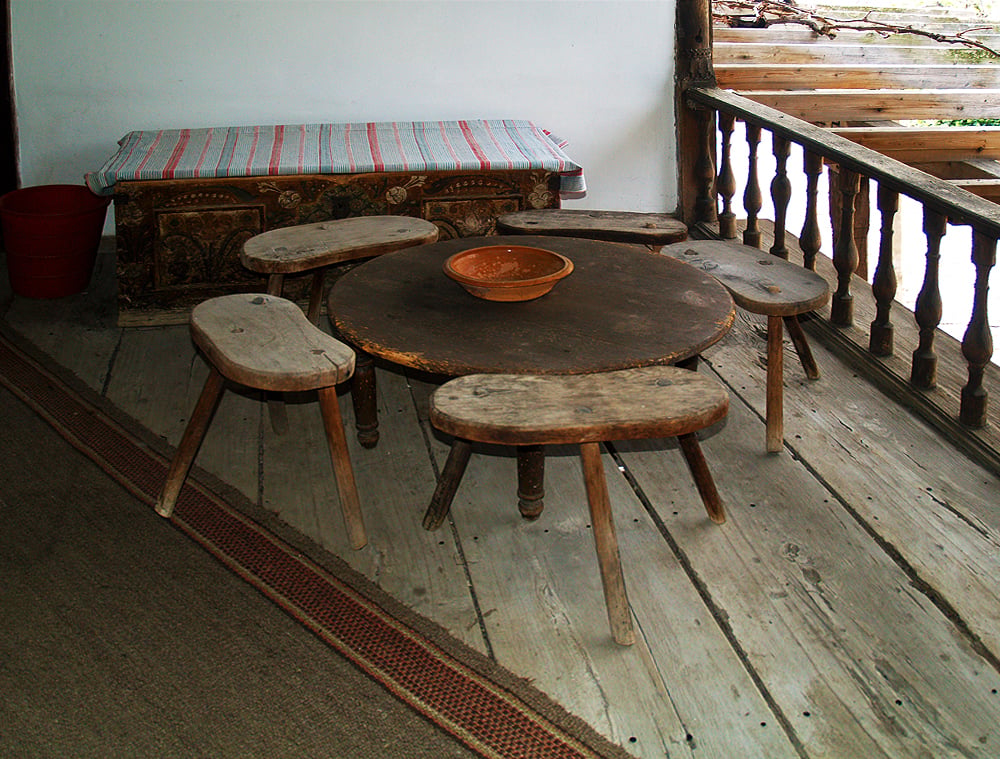 A rustic wooden table with five matching stools is set on a wooden floor. A clay bowl sits in the center. In the background, there's a bench with a striped cloth and a red bucket. The space is enclosed by a wooden railing.
