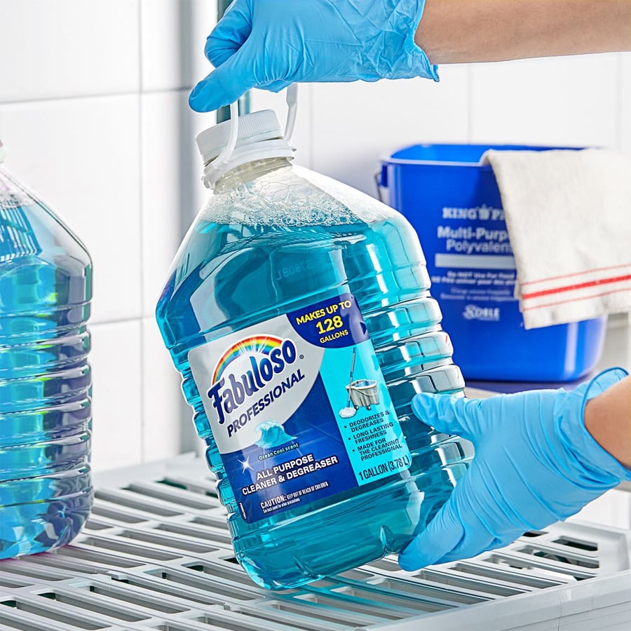 A person wearing blue gloves holds a large jug of Fabuloso Professional all-purpose cleaner in a cleaning supply area with a blue container and towel in the background.