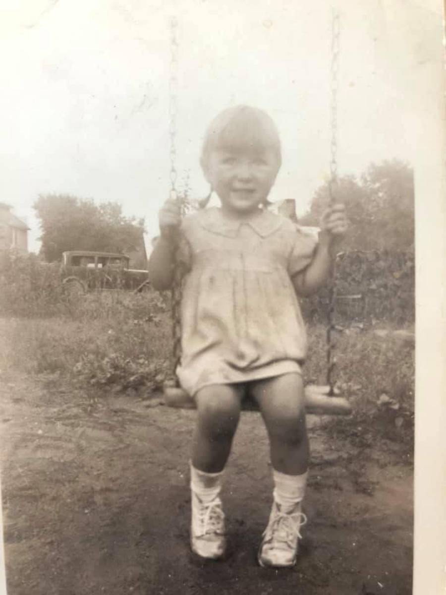 A young child smiles while sitting on a swing outdoors, wearing a light-colored dress, socks, and shoes. The photo is in black and white and appears vintage, with trees and an old car in the background.