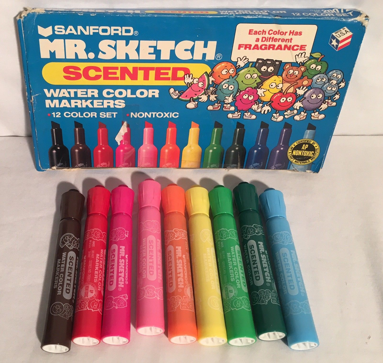 A set of eight Mr. Sketch scented markers in various colors is shown in front of its blue box packaging, which features cartoon marker characters and advertises “Each Color Has a Different Fragrance.”