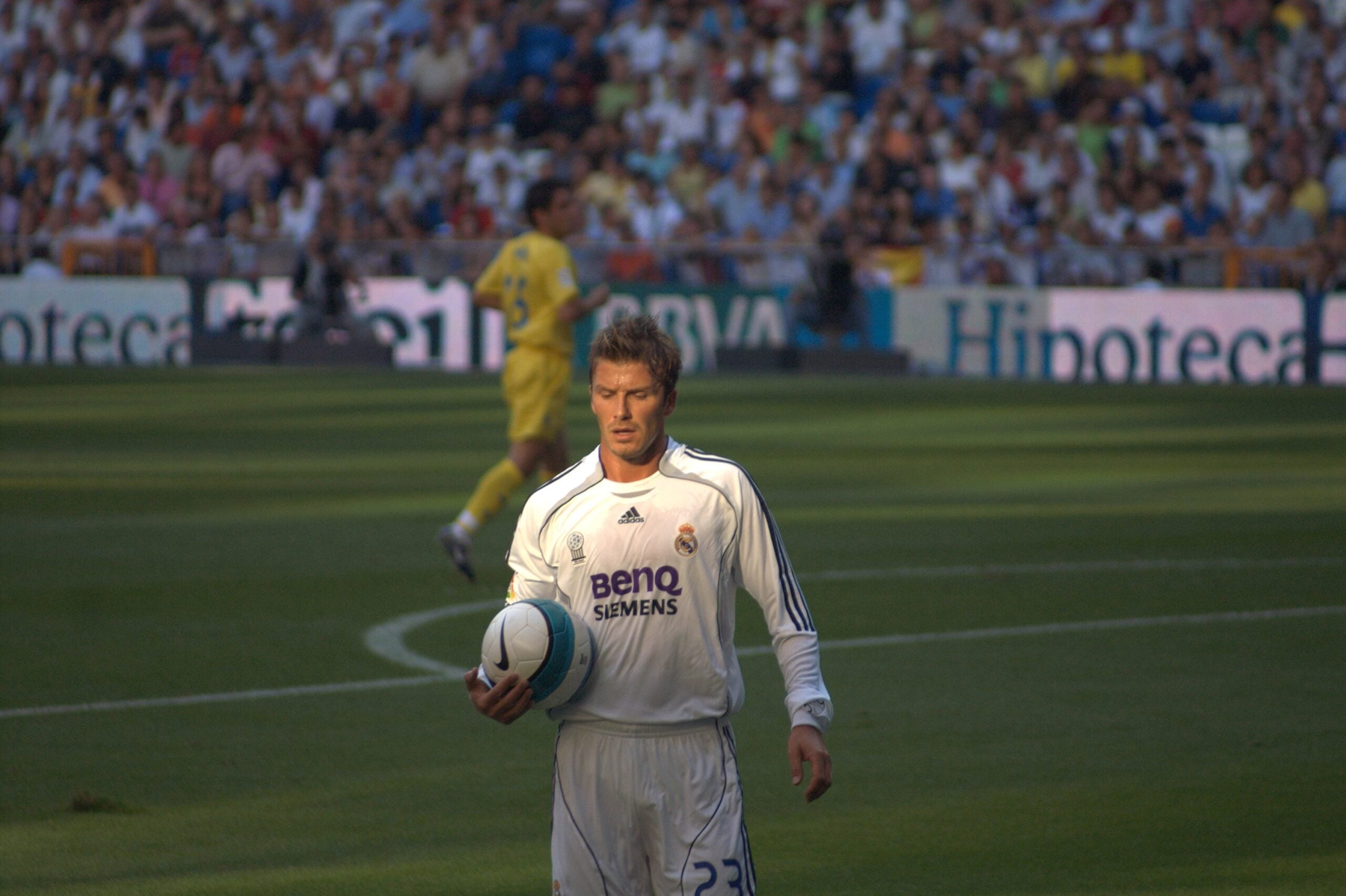 A soccer player in a white Real Madrid uniform holds a ball on a field, preparing for a throw-in. Another player in yellow and a crowd of spectators are visible in the background.