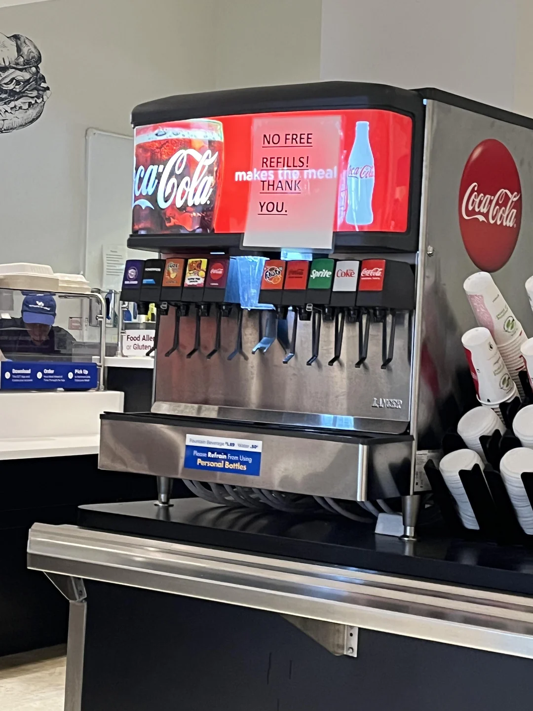 A Coca-Cola soda fountain with various drink options and a red sign on top reading, "NO FREE REFILLS! makes this meal THANK YOU." Cups and lids are on the right side.