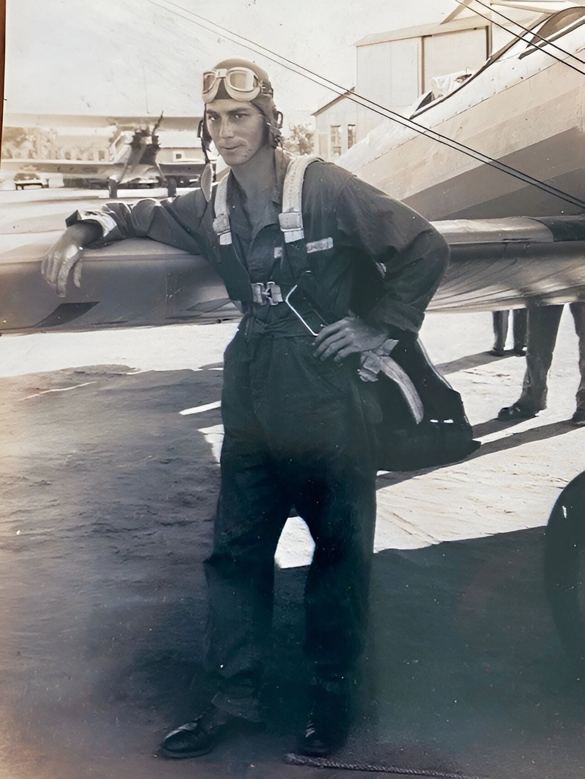 A pilot in a flight suit and goggles leans against a biplane on an airfield. Hangars and another plane are visible in the background. The photo appears vintage, likely from the early or mid-20th century.