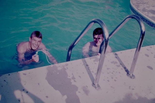 Two people smile while standing in a swimming pool near the metal ladder. One holds a small object. The pool deck is wet, and the water appears blue-green.