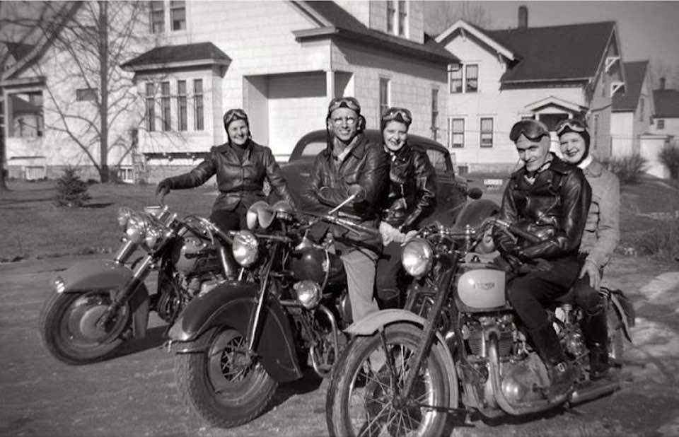 Five people wearing leather jackets and helmets sit on motorcycles, smiling for the camera, with houses and a car visible in the background on a sunny day.