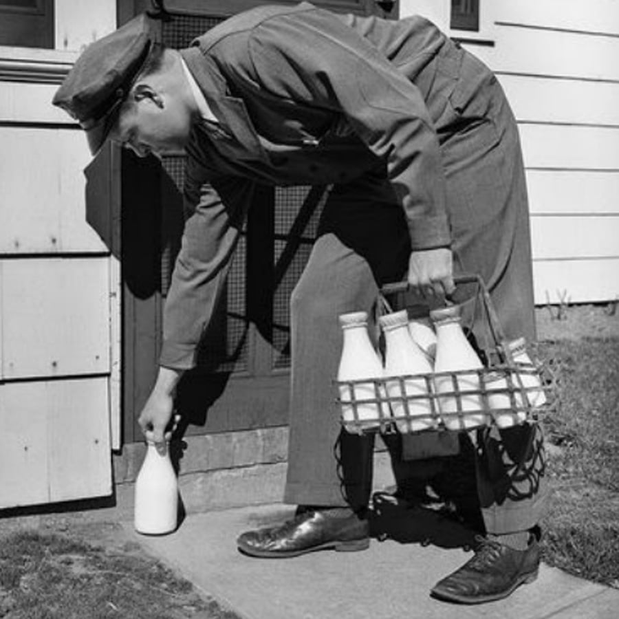 A milkman in a uniform and cap delivers glass bottles of milk to a doorstep, bending down to place one bottle from a wire carrier on the ground beside a house.