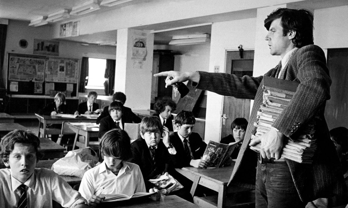 A teacher holding a stack of books points towards students in a classroom. Several students in school uniforms sit at desks, some looking at the teacher, others looking at books. The scene is black and white.