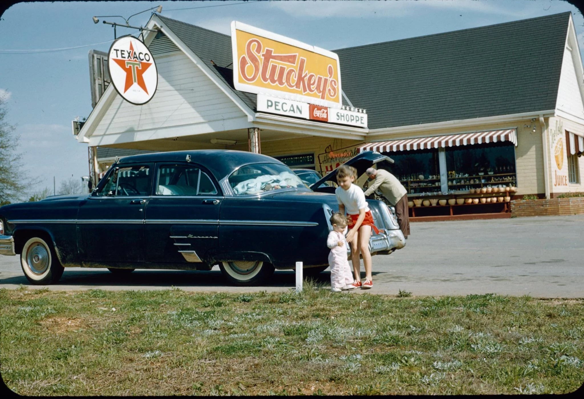 A family unloads luggage from a vintage black car outside a Stuckey’s Pecan Shoppe with a Texaco sign overhead. A child stands beside an adult in front of the shop on a sunny day.