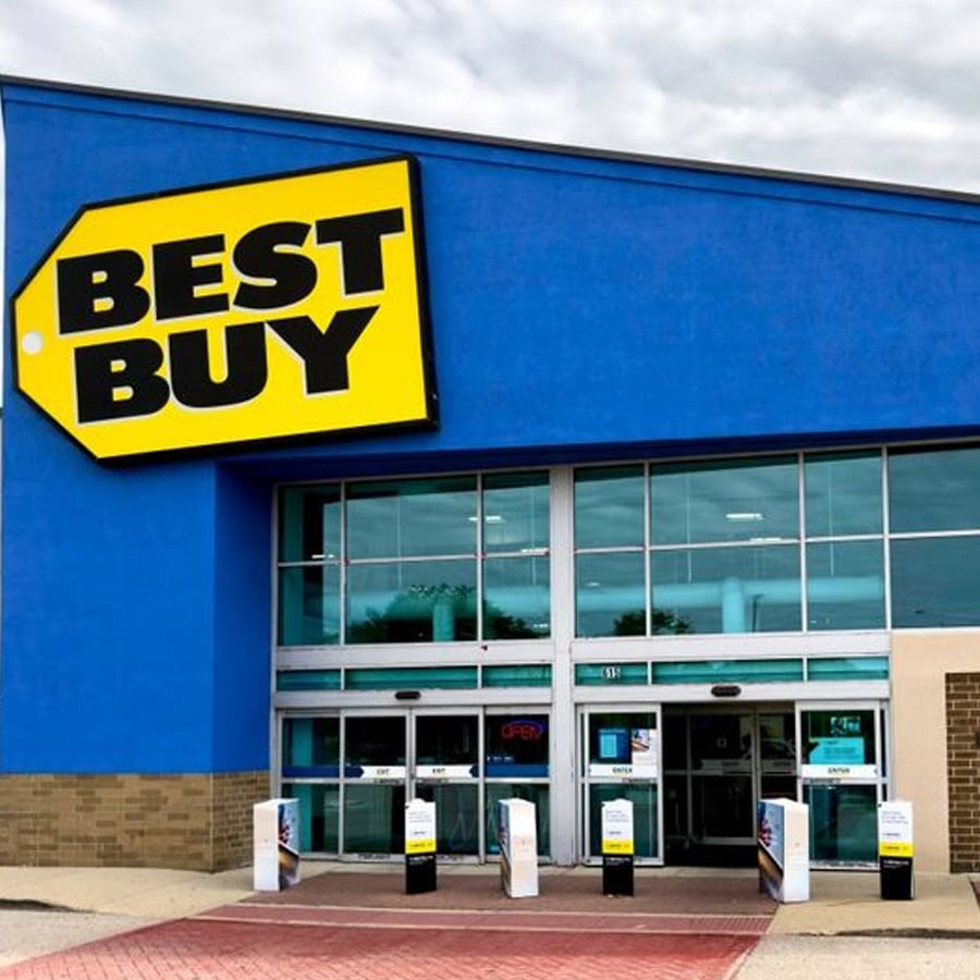 A Best Buy store exterior with a large yellow sign, automatic glass doors, and a blue facade under a cloudy sky.