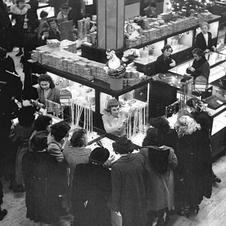 Black and white photo of women crowded around a jewelry counter in a busy department store, with one clerk assisting customers. Holiday decorations and displays are visible in the background.